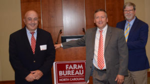 Administrators from NC State University's College of Agriculture and Life Sciences stand by a podium while smiling. From left to right the faculty include Rich Bonanno, associate dean and director of NC State Extension; Richard Linton, dean of the College of Agriculture and Life Sciences; and Steve Lommel, associate dean and director of the North Carolina Agricultural Research Service.