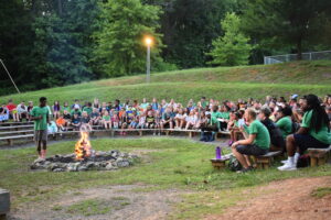 About 100 youth sitting on bleachers around a campfire watching a man talk