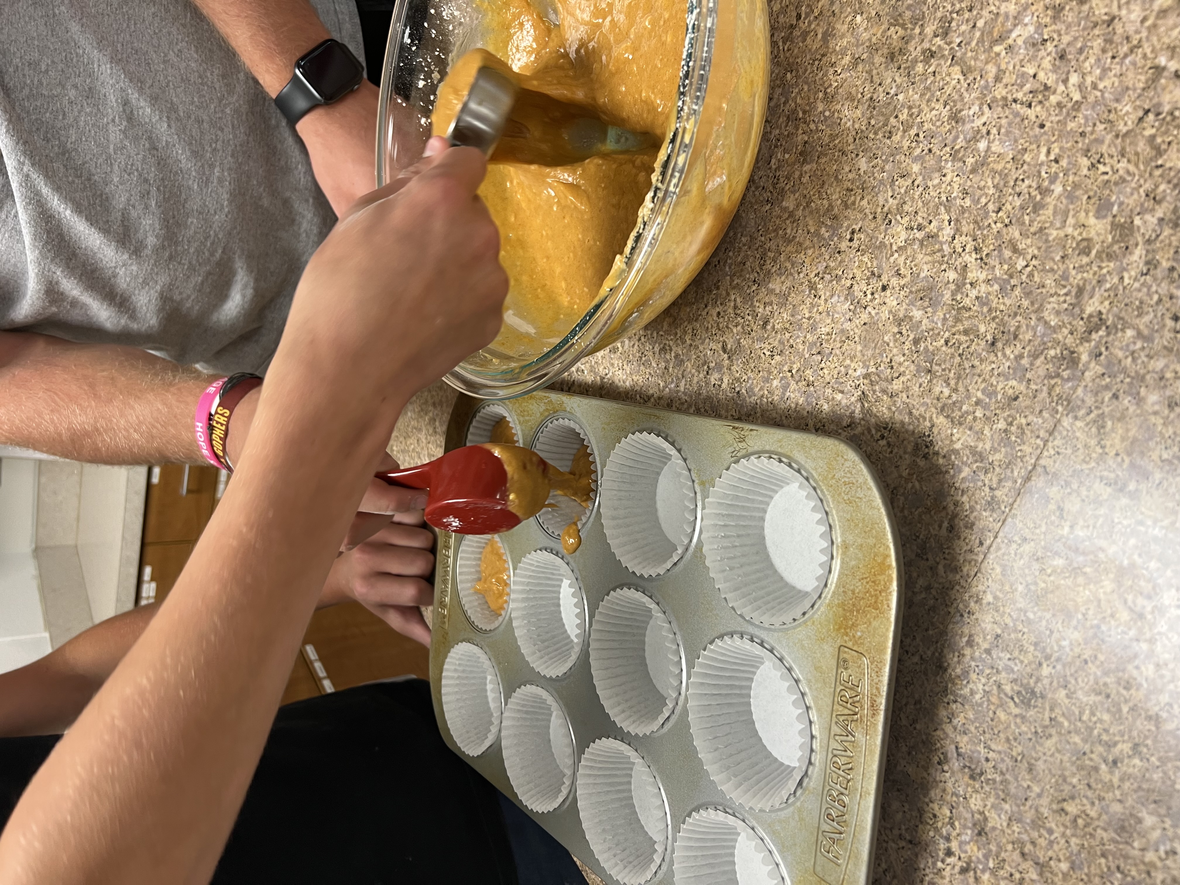 Two students use measuring cups to fill empty muffin tins with batter.