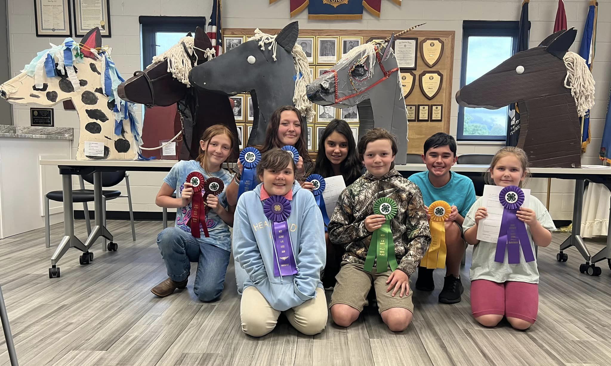 A group of kids pose with ribbons in front of crafted horse heads.