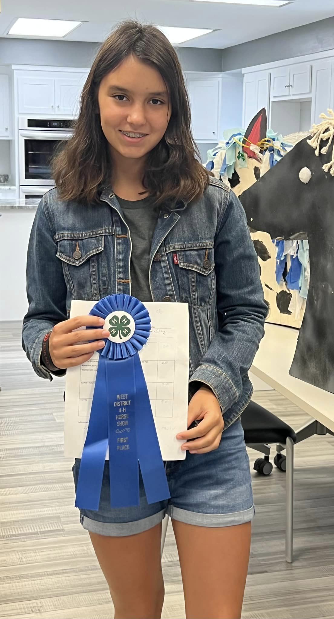 A girl poses with a blue ribbon. Crafts are on the table behind her.