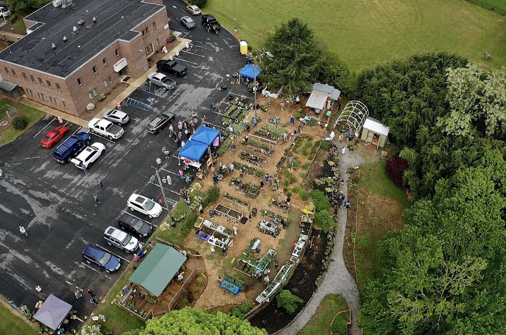 An aerial view of the entire plant sale event, showing the garden area, vendor tables, greenhouse, and parking lot filled with cars and shoppers. The sale is set up on a landscaped plot with walking paths, raised garden beds, and clusters of plants for sale, surrounded by trees and open green space.